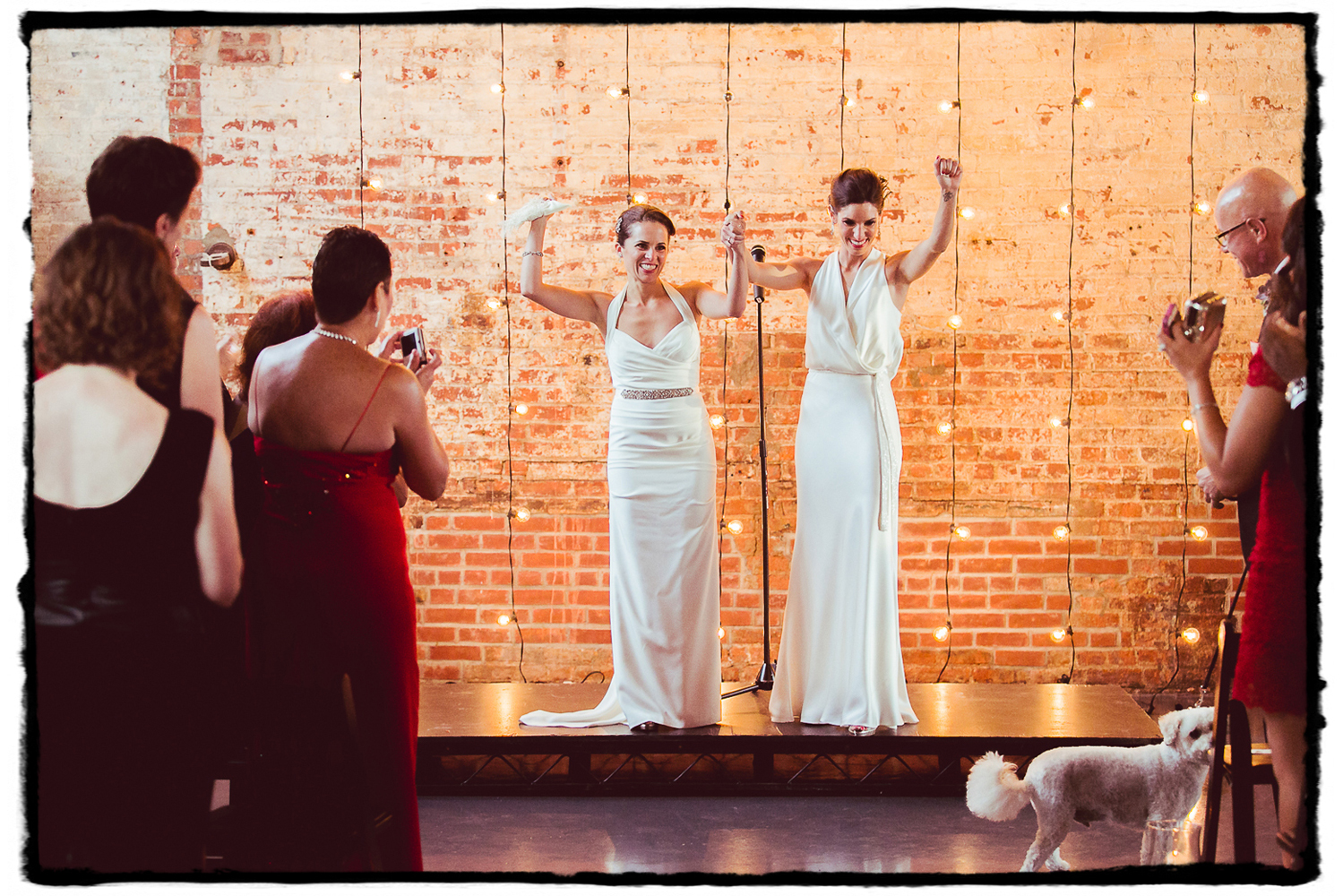 Lara & Nicole were married at The Green Building in front of a brick wall illuminated by hanging string lights.